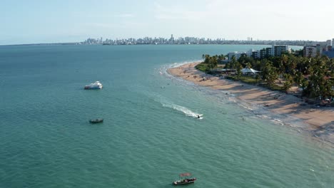 tilt up aerial drone shot of the tropical bessa beach in the capital city of joao pessoa, paraiba, brazil with people enjoying the ocean, a small tour boat, jet ski, and skyscrapers in the background