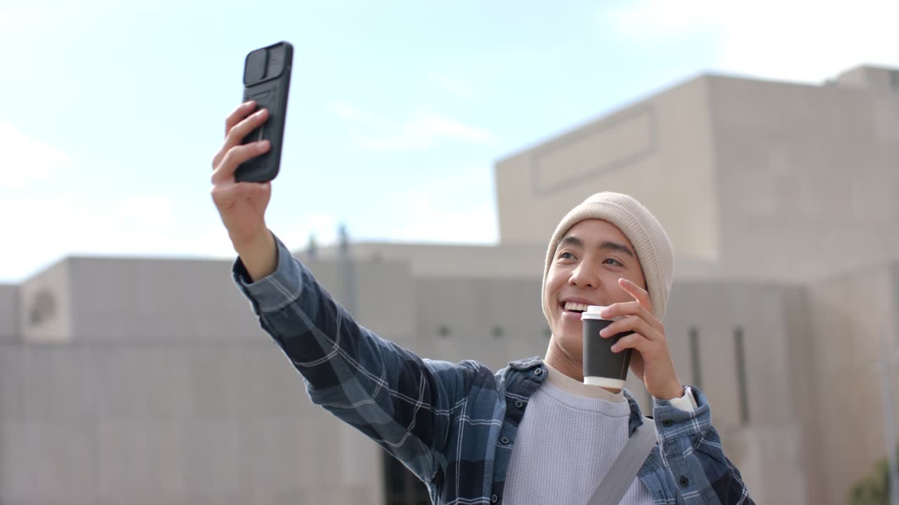 Asiático segurando um smartphone no terraço para uma chamada ao vivo, tomando café e assentindo para a tela