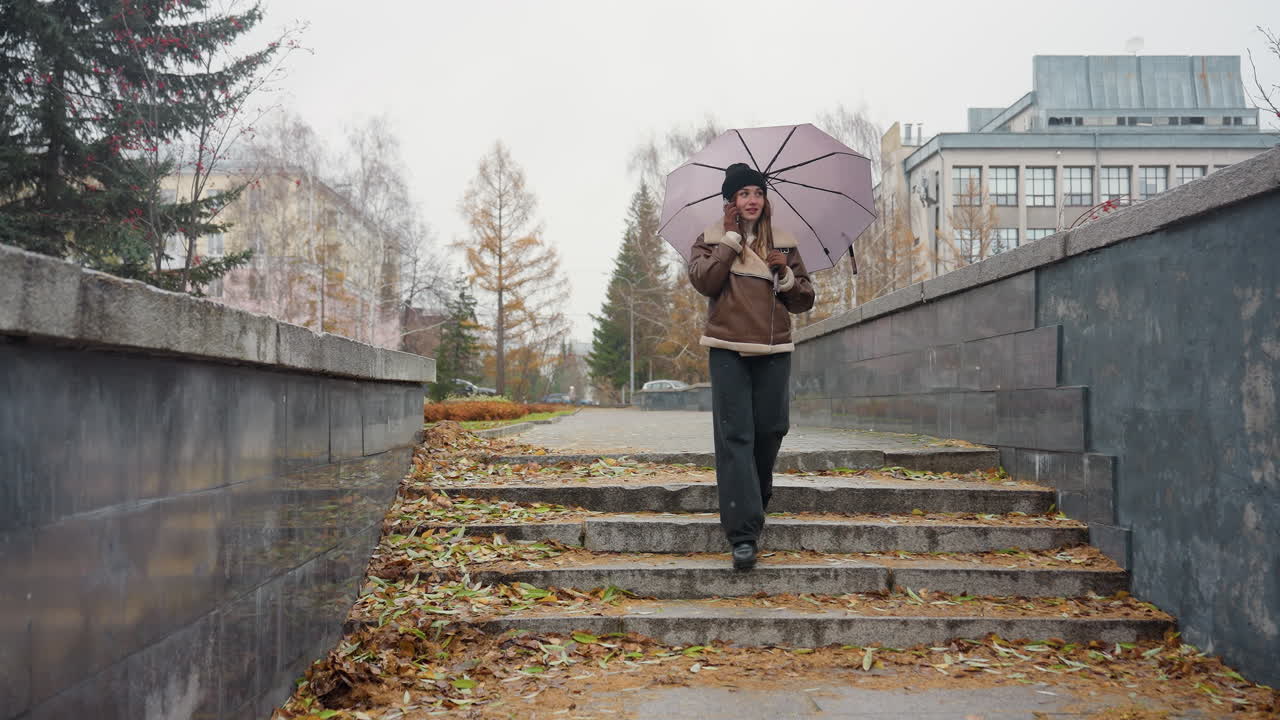 Young girl smiling, making phone call while holding umbrella, wearing black knit cap, brown shearling jacket, black trousers, walking through urban park on cold overcast day with light snowfall