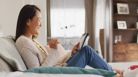 Video-of-happy-asian-woman-resting-on-sofa-with-tablet