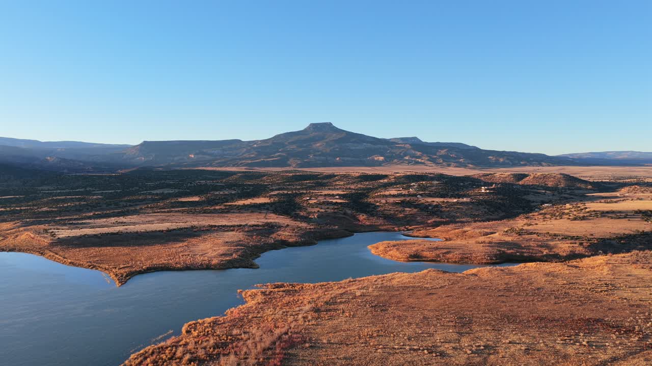 Aerial view captures the blue reservoir water of Abiquiu Lake below the volcanic basalt peak of Cerro Pedernal. Arid brush and sunlit shorelines define the high desert plateau of northern New Mexico