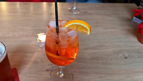 Cocktail-closeup-on-wooden-table,-savory-drink-merged-with-an-orange-slice-in-fresh-ice