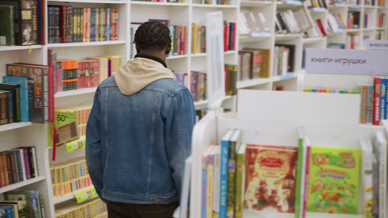 Back view of person in denim jacket with hoodie walking between colorful bookshelves in bookstore. Shelves filled with books in various genres, highlighting vibrant book spines