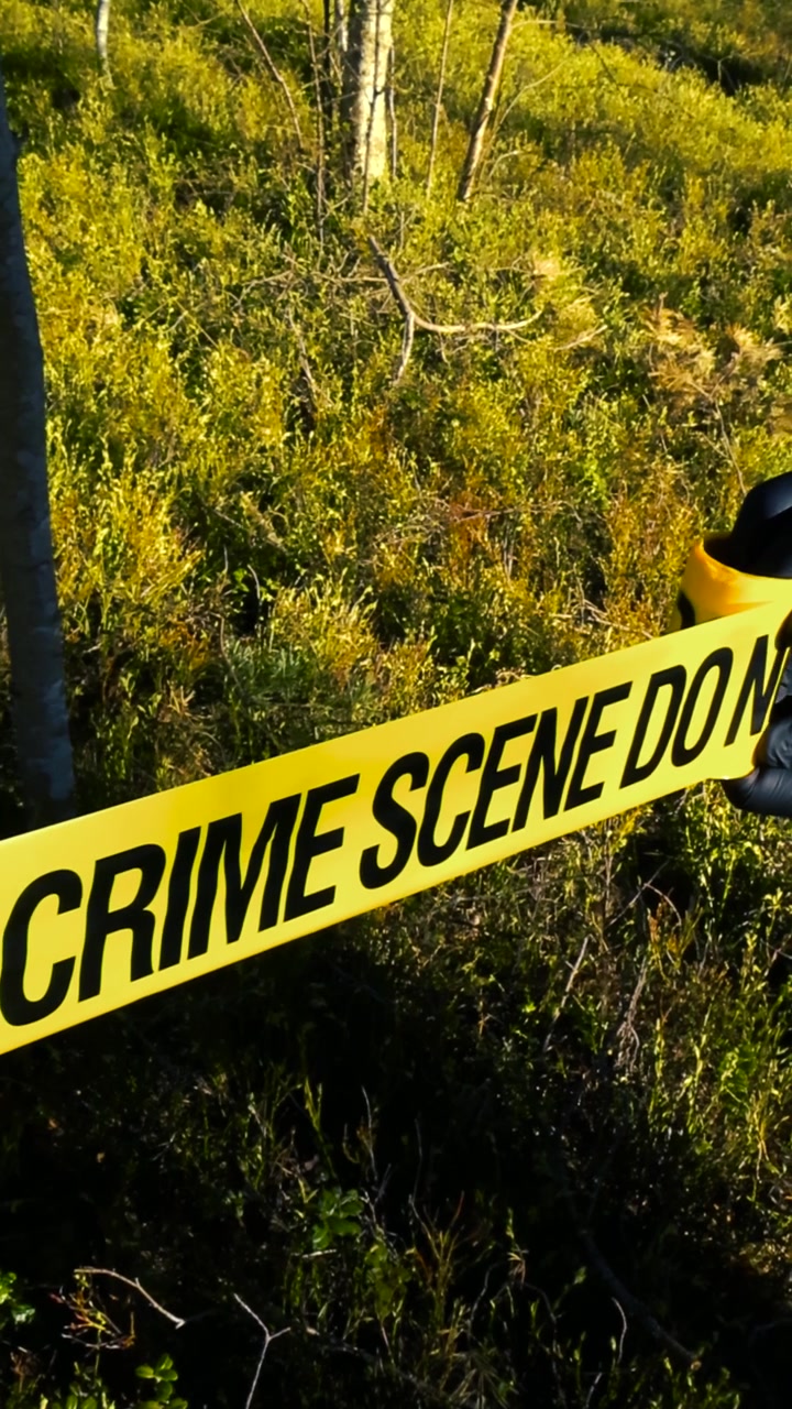 Gloved hands of a law enforcement officer or detective unrolling yellow Crime Scene Do Not Cross tape, securing an investigation area in a sunlit forest during a homicide investigation
