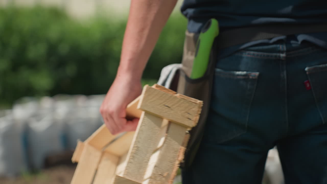 Close up rear view of farmer walking toward farm carrying wooden crate with tool belt visible on hip, sunlit garden beds and greenhouse frame blurred in background