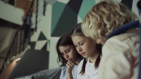 Two-schoolgirls-looking-down-sitting-modern-hallway.-Girls-watching-tablet.