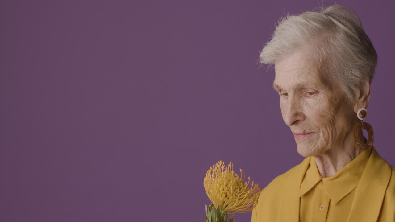 Side View Of Mature Woman With Short Hair Wearing Mustard Colored Shirt And Jacket And Earrings Posing Holding A Flower On Purple Background