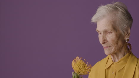 Vista-Lateral-De-Una-Mujer-Madura-Con-El-Pelo-Corto-Usando-Camisa-Y-Chaqueta-De-Color-Mostaza-Y-Aretes-Posando-Sosteniendo-Una-Flor-Sobre-Fondo-Morado