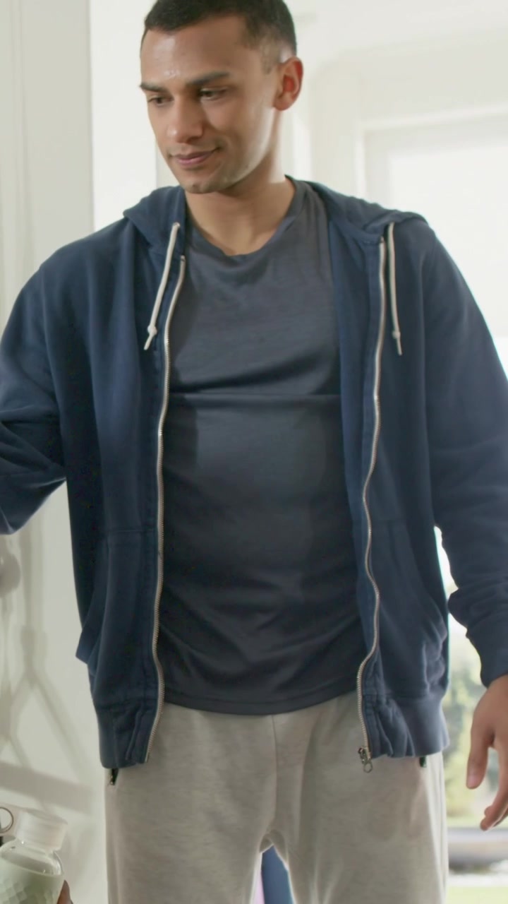 Vertical video: Male in navy hoodie accepting pale green bottle at home, offered by friend to drink