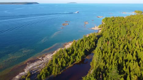 Aerial-drone-shot-following-the-beautiful-coastline-of-Georgian-Bay-as-a-boat-heads-out-into-the-vast-blue-ocean-waters,-Ontario,-Canada