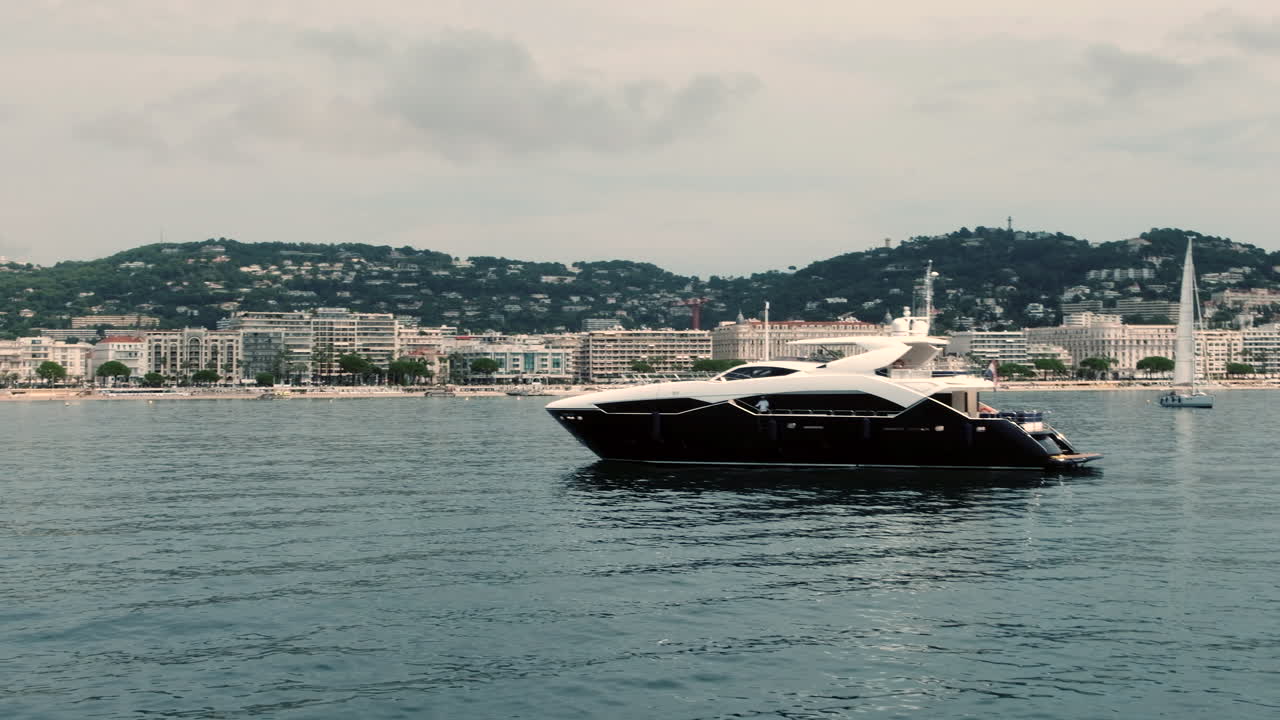 Un elegante yacht in bianco e nero scivola sul mare calmo vicino alla costa della Costa Azzurra, con edifici moderni e barche a vela sullo sfondo.