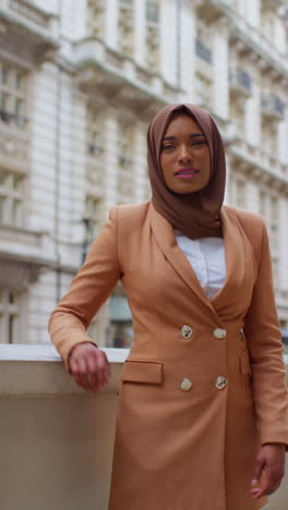 retrato vertical de una mujer de negocios musulmana sonriente con hijab y traje de negocios moderno de pie fuera de los edificios de oficinas de la ciudad