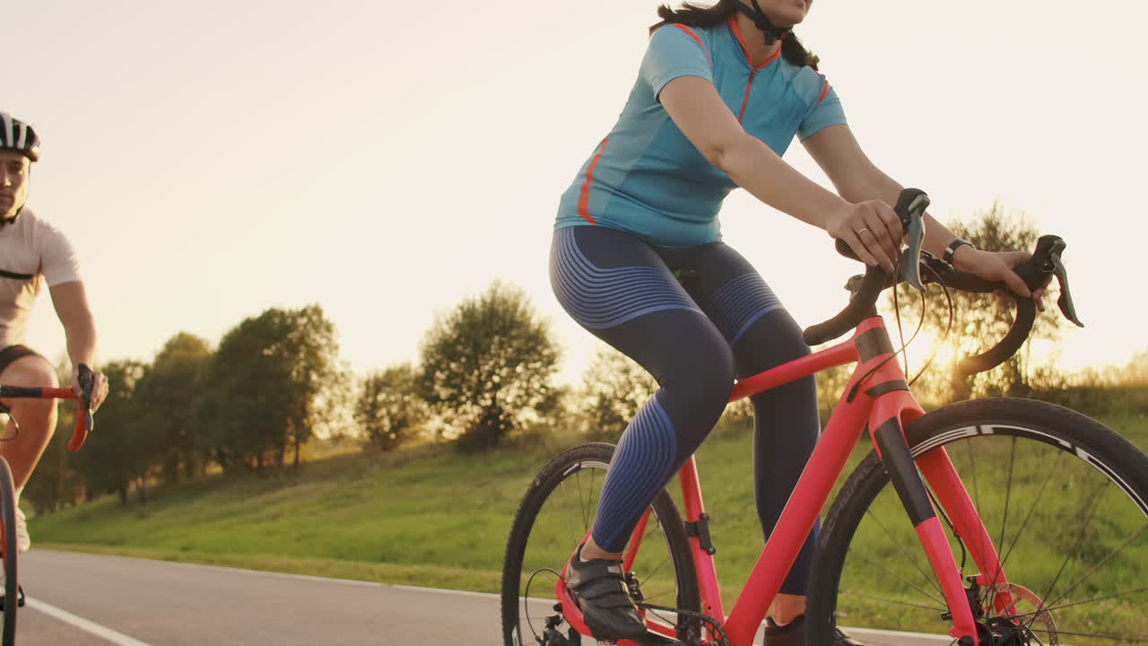 두 명의 자전거 타는 남자와 여자가 서서히 움직이는 해가 지는 동안 헬과 스포츠 의류를 입은 도로 자전거를 타고 고속도로를 타고 있습니다.