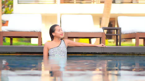 Joven-Asiática-Descansa-Dentro-Del-Agua-Al-Borde-De-La-Piscina-Junto-A-La-Línea-De-Tumbonas-En-El-Complejo-Hotelero-Sunset,-En-Cámara-Lenta