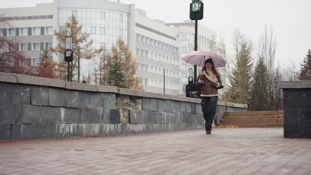 Female runner in light snowfall holding umbrella, wearing knit cap, brown shearling jacket, black trousers, one hand in pocket, enjoying winter day with autumn leaves scattered on ground