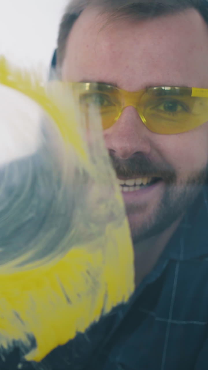 Dolly cam: smiling constructor with goggles removes bright yellow paint from glass with rag at construction in empty room