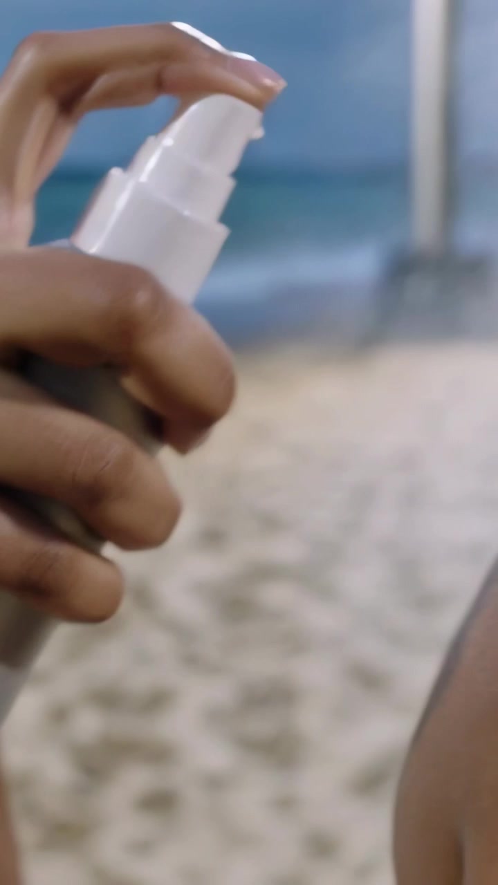 Vertical video: Spraying white bottle, swimsuit pal rubbing shoulder for sun on beach, copy space