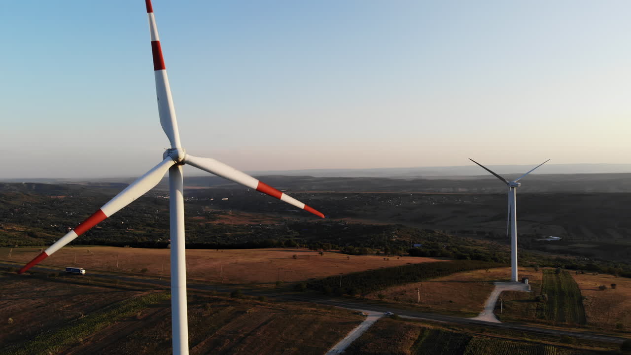 Aerial view of tall wind turbines turning gently at sunset, surrounded by wide open fields and distant hills