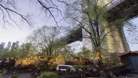 una perspectiva única desde debajo del puente de brooklyn, mirando hacia arriba a través de los árboles con luces de cuerda, capturando la estructura del puente contra un cielo claro de la noche