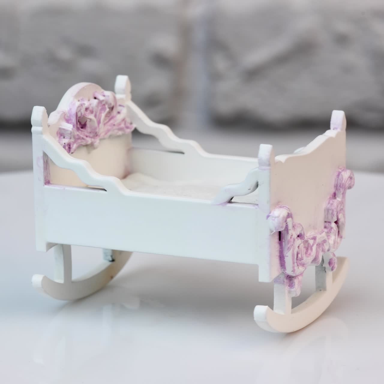 Tiny baby cradle at the rotating table. White toy crib with carved ornaments shaded pink. Close up. White wall background