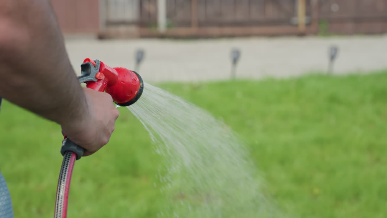 close up of hairy hand holding red garden nozzle watering green grass in backyard, water spray streams evenly across lawn, capturing daily outdoor care routine in bright daylight with vivid clarity