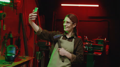 Caucasian-welder-woman-in-goggles-and-holding-hammer-making-a-selfie-with-smartphone-in-metal-workshop
