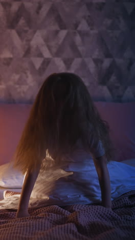 active little child with flying blonde hair has fun on soft bed near high play wigwam with glowing night lamp inside in dark bedroom at home slow motion