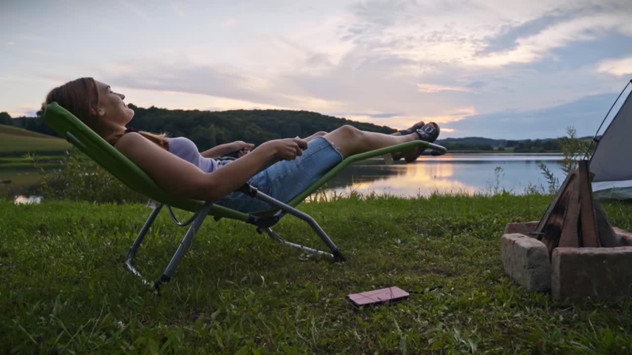 Kvinde ligger på udendørs stol nær sø og bål, kaster smartphone til side efter brug, hvilket viser digital detox-konceptet og en fredelig campinglivsstil i Savci, Slovenien.