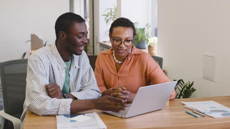 Junger-Arbeitnehmer,-Der-Mit-Einem-Laptop-Arbeitet,-Der-An-Seinem-Schreibtisch-Sitzt,-Während-Die-Geschäftsfrau-Mit-Ihm-Spricht,-Während-Er-Am-Schreibtisch-Sitzt,-Während-Sie-Auf-Den-Laptop-Schaut