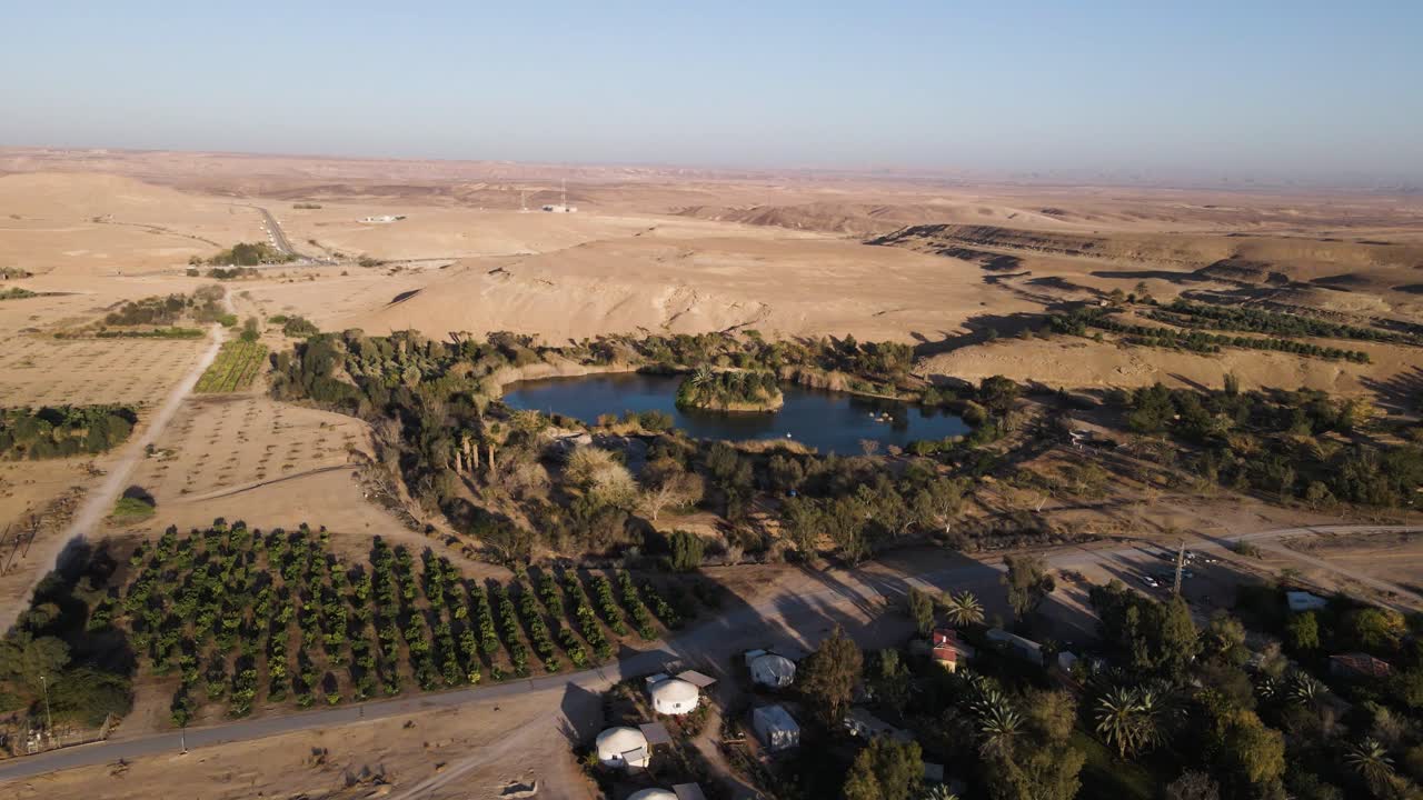 Drone push-in over Neot Smadar oasis in Israeli desert, aerial view, lush greenery, central water body and vast arid surroundings.