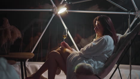 upset young woman drinks alcoholic drink in glass sitting in armchair at night in glamping hotel room. unpleasant feeling of loneliness and devastation