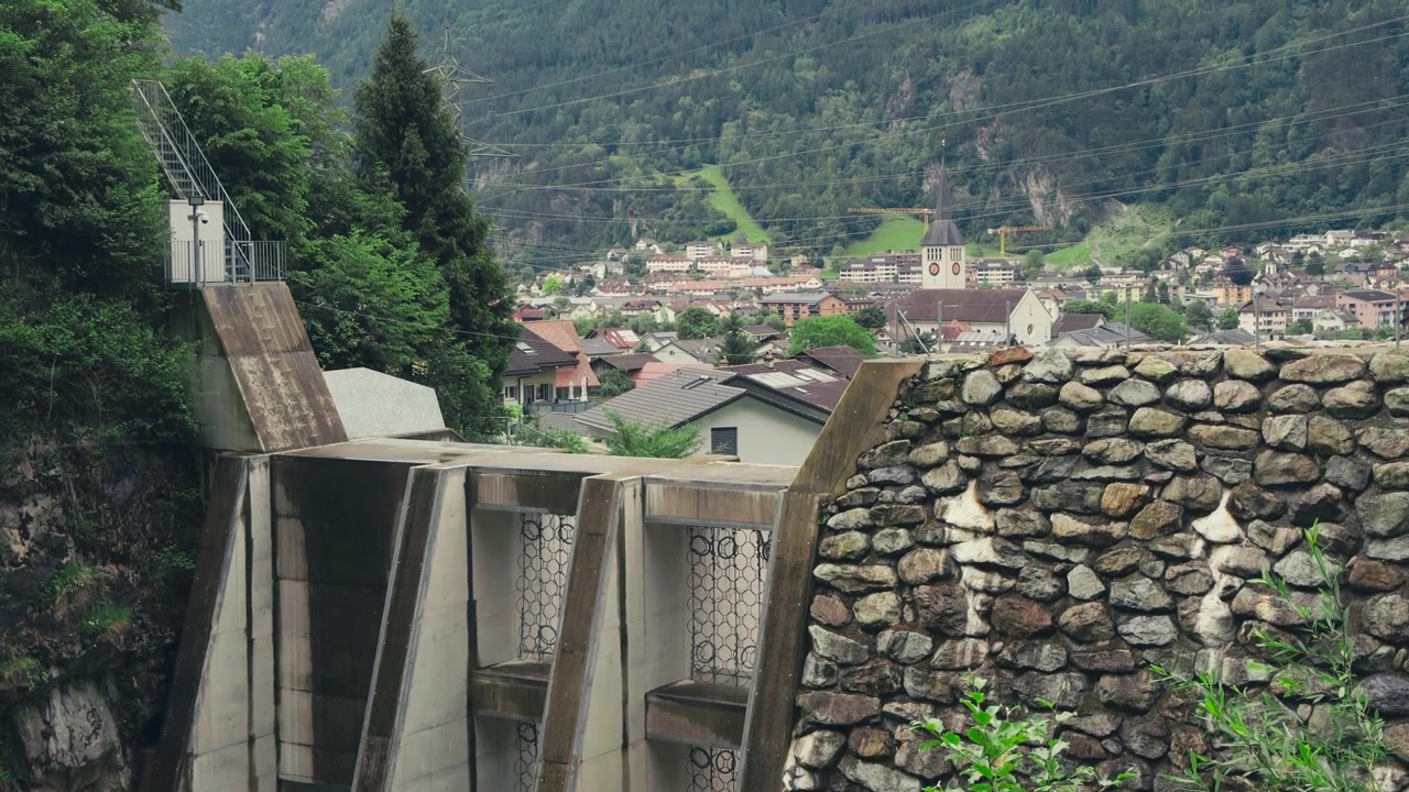 Imagem estática de uma comporta de usina hidrelétrica nas montanhas com a vila alpina protegida ao fundo, destacando a gestão da água, o controle de enchentes e a infraestrutura crítica