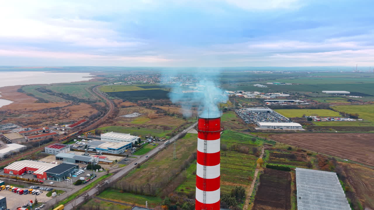 Vista aerea di una ciminiera di fabbrica che emette fumo sui campi. Ciminiera di centrale termica con fumo blu situata tra campi agricoli e una zona industriale.