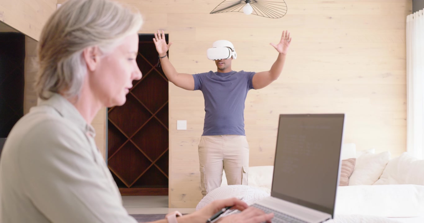 Casal diverso em casa, homem colocando óculos de VR branco explorando o mundo enquanto a mulher digita no laptop.