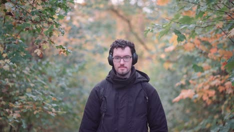 Joyful-bearded-European-man-with-trendy-glasses-stands-amidst-the-hues-of-a-mixed-autumn-forest,-radiating-happiness-and-warmth-upon-seeing-viewers,-emanating-pure-positive-vibes