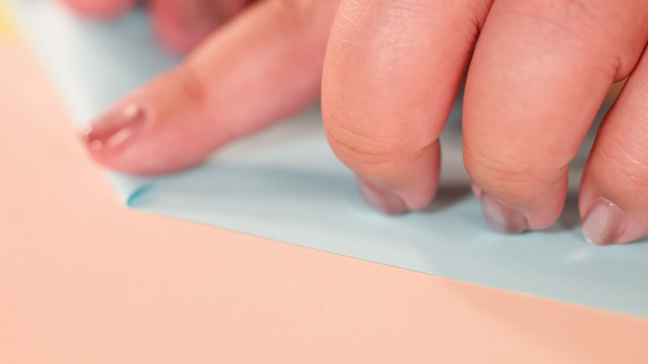Primer plano de unos dedos doblando cuidadosamente un papel azul sobre una superficie color melocotón con iluminación suave.