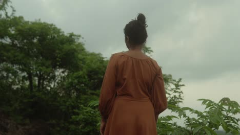 a woman in an orange dress stands on a hillside looking out into nature under a cloudy sky