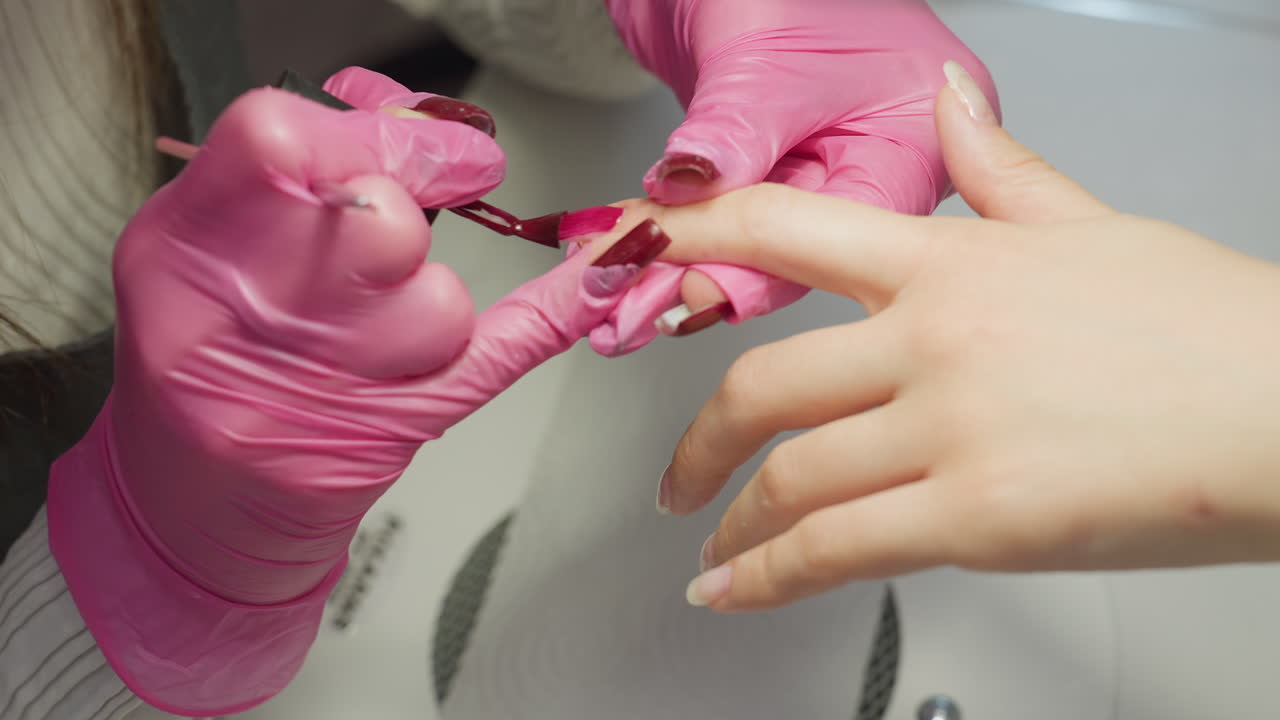 Close up of nail technician wearing pink gloves carefully applying red nail polish to client finger with precision, highlighting manicure process and steady hand control in salon environment