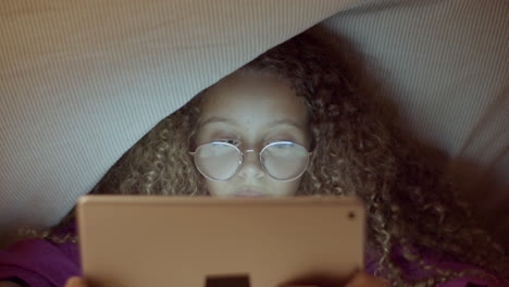 Front-view-closeup-of-cute-girl-reading-ebook-under-blanket