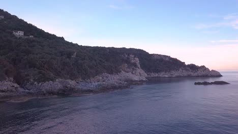 Forward-and-crane-up-aerial-shot-over-calm-sea-and-next-to-a-rocky-shore-during-blue-hour-in-Greece