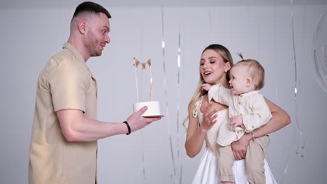 caucasian mother wearing white dress holding a cute baby in hands. father brings a cake and child stretches hands to it. studio footage.