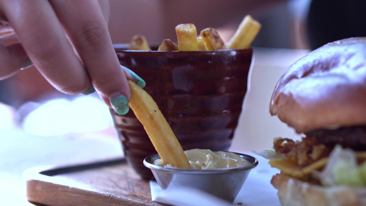 Close up of female hand taking a piece of delicious fries from bowl full of fried potatoes with burger in foreground. Dipping fries in mayonnaise.
