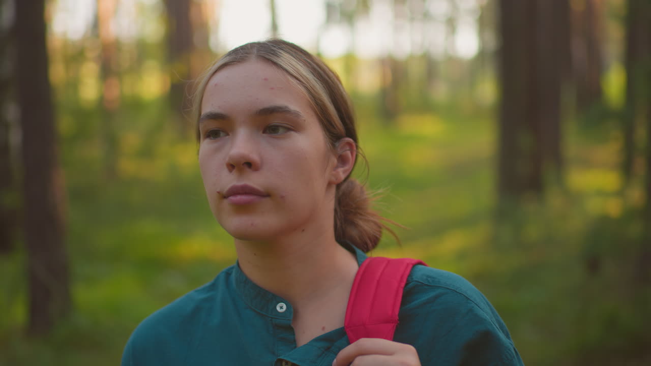 primer plano de una mujer caminando por un bosque tranquilo, ajustando la correa de la bolsa roja en el hombro, mirando pensativo en la distancia con la cálida luz del sol proyectando un suave resplandor en el rostro
