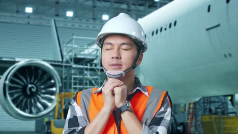 cercano de ingeniero masculino asiático con casco de seguridad reza por algo mientras está de pie con aviones en el hangar