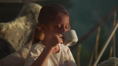 kid drinks warm milk in evening. sleepy schoolgirl looks at table thoughtfully feeling tired after active day on blurred background slow motion