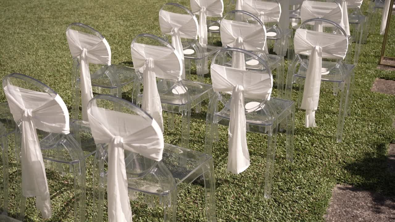 Transparent chairs with white fabric bows elegantly arranged outdoors on a grassy surface for a wedding ceremony