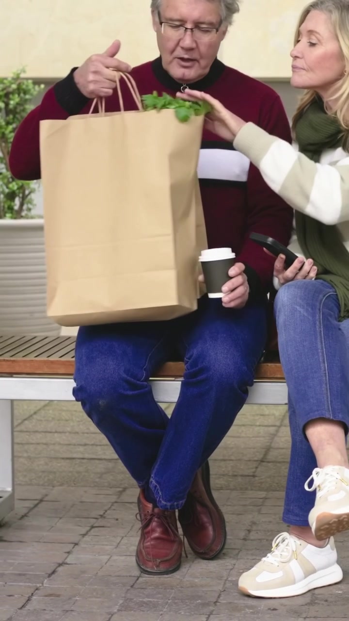 Video verticale: Coppia che parla seduta su una panchina di legno, l'uomo tiene una tazza e solleva una borsa mostrando verdure.