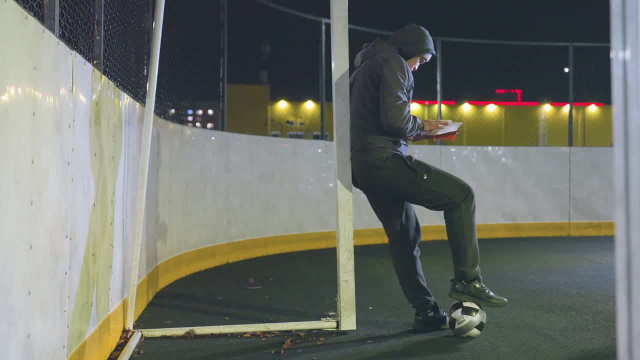 hombre con capucha apoyado en el poste de la portería al aire libre por la noche, leyendo un libro con el pie derecho en la pelota de fútbol, luces urbanas brillando en el fondo, movimientos sutiles de follaje en el suelo