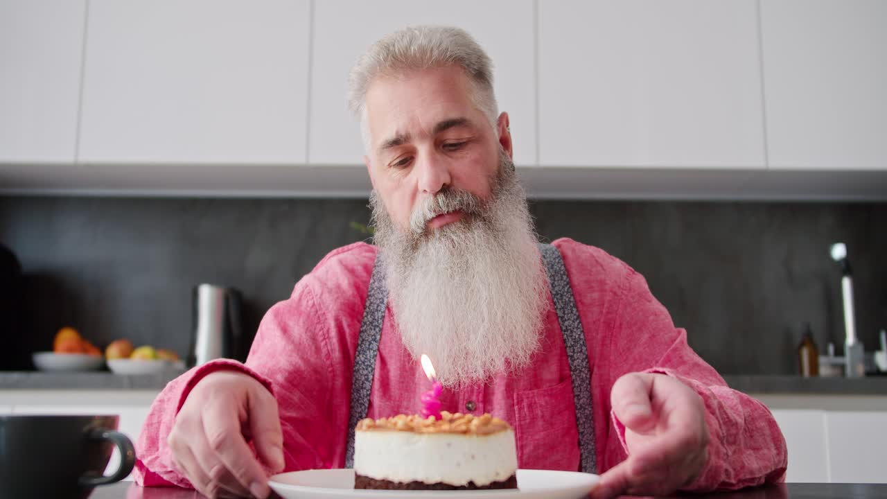 A sad and lonely man with gray hair with a lush beard in a pink shirt looks at a cake on which one candle is burning during his birthday and blows it out in a modern apartment