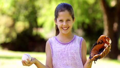 Sporty-little-girl-smiling-at-camera-in-the-park-playing-with-baseball